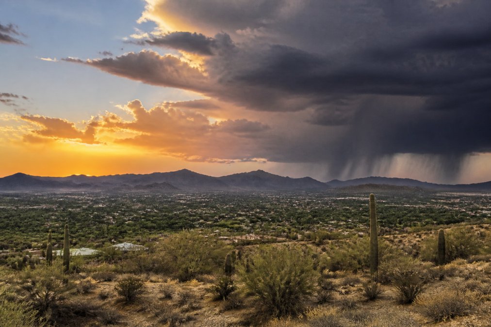 Desert landscape at sunset with dramatic monsoon storm clouds gathering over distant Phoenix area mountains, showing contrast between clear skies and dark approaching weather system
