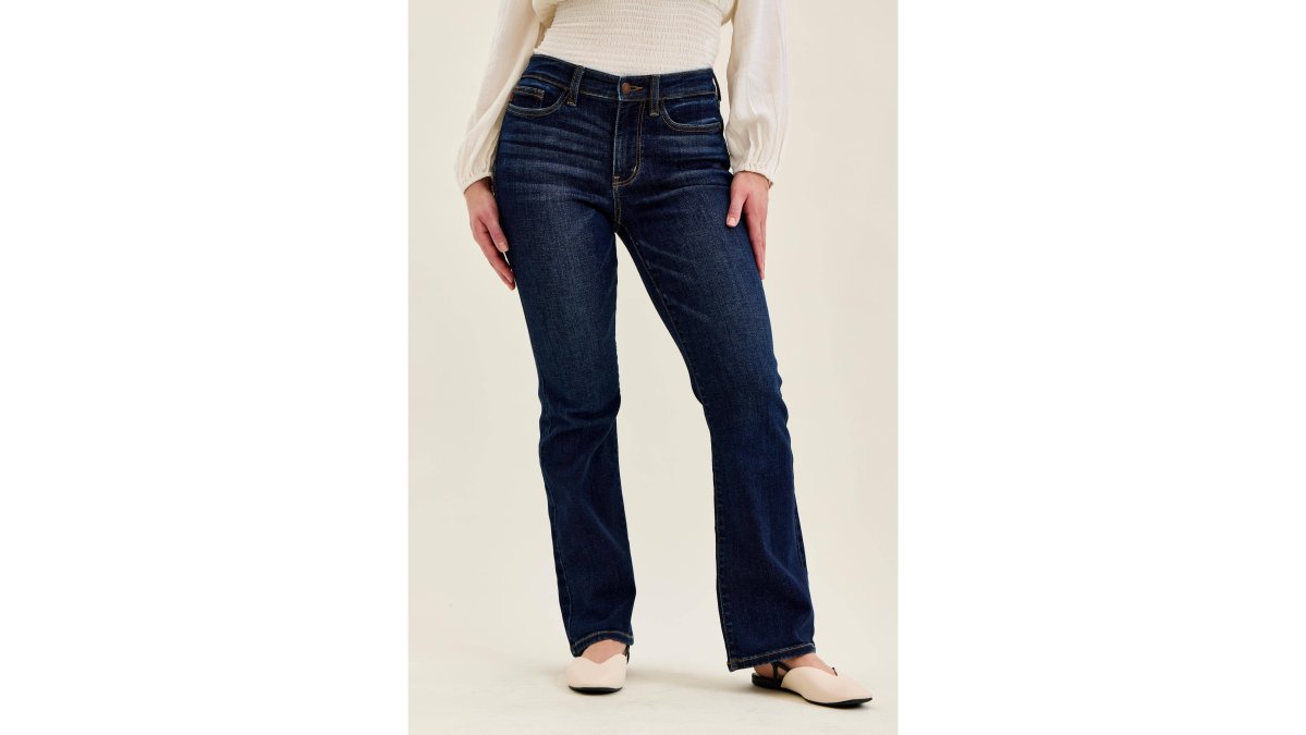 A petite woman around 5'4" standing in a bright minimal boutique, wearing dark wash bootcut jeans and a fitted white top, photographed from the side to show the jean length sitting cleanly at the floor and the fit through the hip and thigh