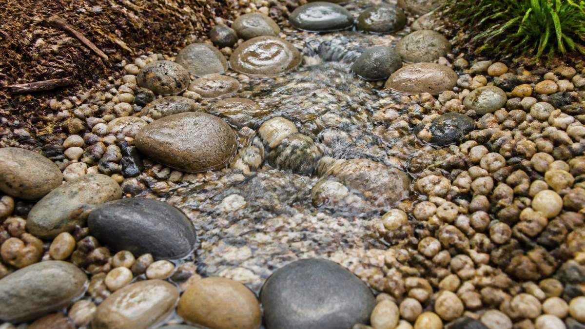 Close-up of smooth river rock and pea gravel with water flowing through the stones in a landscaped drainage area, showing water permeability in action