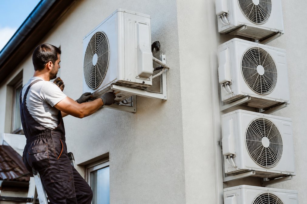 Professional HVAC technician in work clothes mounting an outdoor air conditioning unit on a building wall using tools, multi split system visible nearby, natural daylight