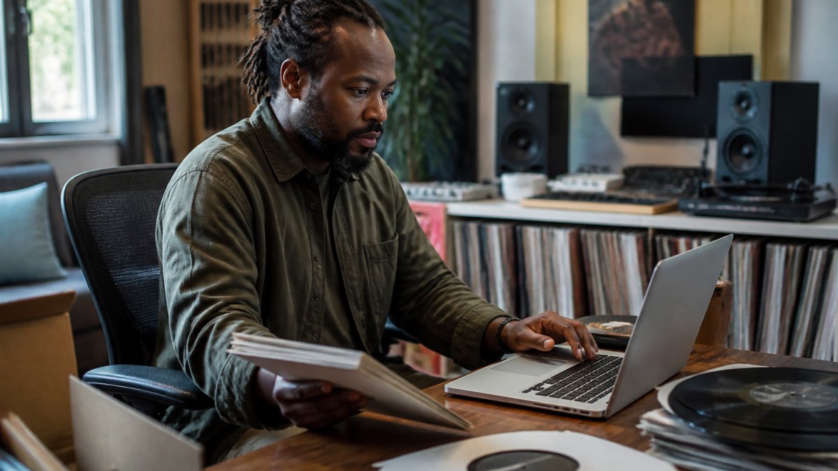 A music label manager reviewing artist rosters and release schedules on a laptop in a modern home studio, with vinyl records and studio monitors visible in the background