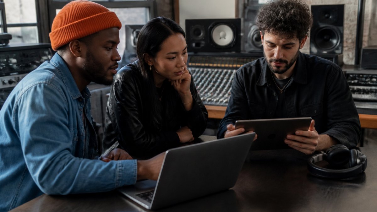 A music label team reviewing release analytics on a laptop and tablet in a modern studio office, with headphones and a mixing console visible in the background