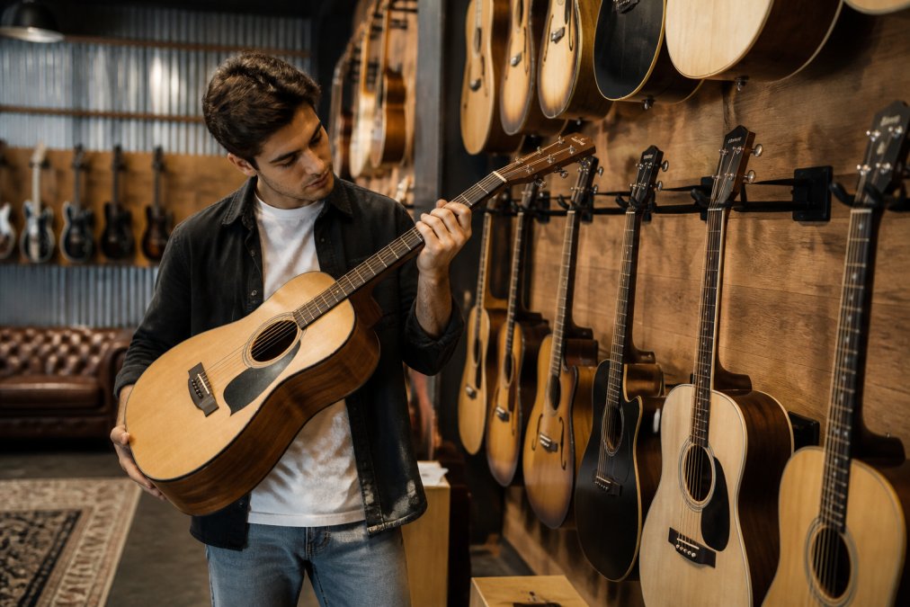 A musician browsing guitars on display in a well-lit music store, examining the neck of an acoustic guitar with shelves of instruments in the background