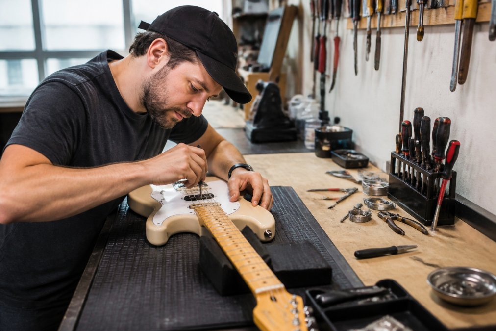 A guitar technician adjusting the truss rod on an electric guitar in a bright workshop, with tools and spare parts neatly arranged on the bench