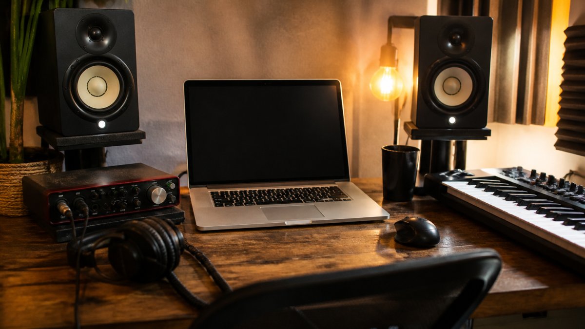A home studio desk with a laptop displaying a DAW multitrack session, studio monitors, and an audio interface, with warm lighting and a MIDI keyboard nearby