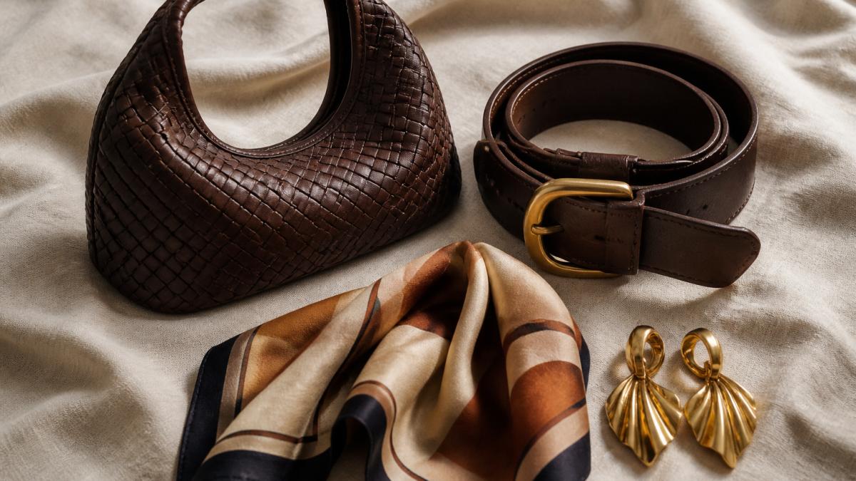 Flat-lay photograph on a neutral linen surface showing a woven leather handbag, an oversized leather belt coiled beside it, a folded triangle scarf, and a pair of gold statement earrings arranged together as a styled accessory set, soft natural window light