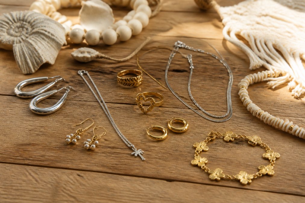 Verzameling van verschillende sieradenstijlen uitgespreid op een houten tafel met warm natuurlijk licht: zilveren oorbellen, gouden ringen, roestvrij stalen kettingen en armbanden in verschillende texturen