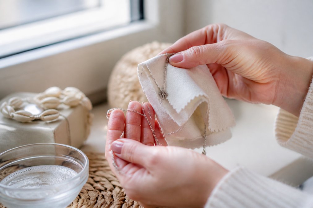 Close-up van voorzichtige handen die een zilveren ketting schoonmaken met een zachte microfiber doek, met een klein bakje met zilvercleaner en een natuurlijke achtergrondverlichting van een raam