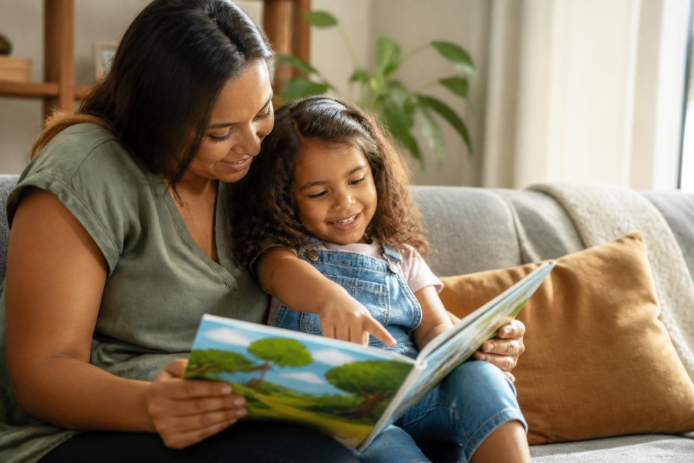 A parent and young child cuddled on a couch reading a colorful picture book together, warm natural light filling the living room, child smiling and pointing at an illustration