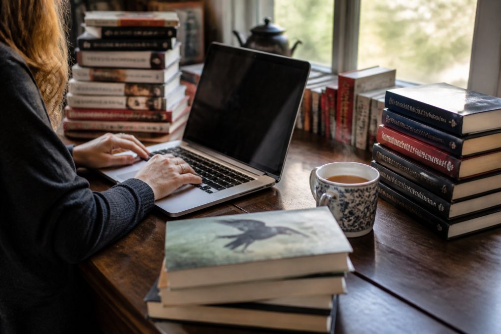 A person browsing mystery and crime fiction titles on a laptop at a wooden desk, with stacks of physical books nearby and a warm cup of tea steaming beside the keyboard, natural afternoon light from a window