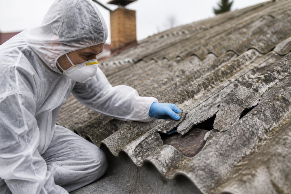 Zblízka poškozená eternitová střecha s viditelným odlupováním materiálu, pracovník v ochranném obleku a respirátorem provádějící inspekci