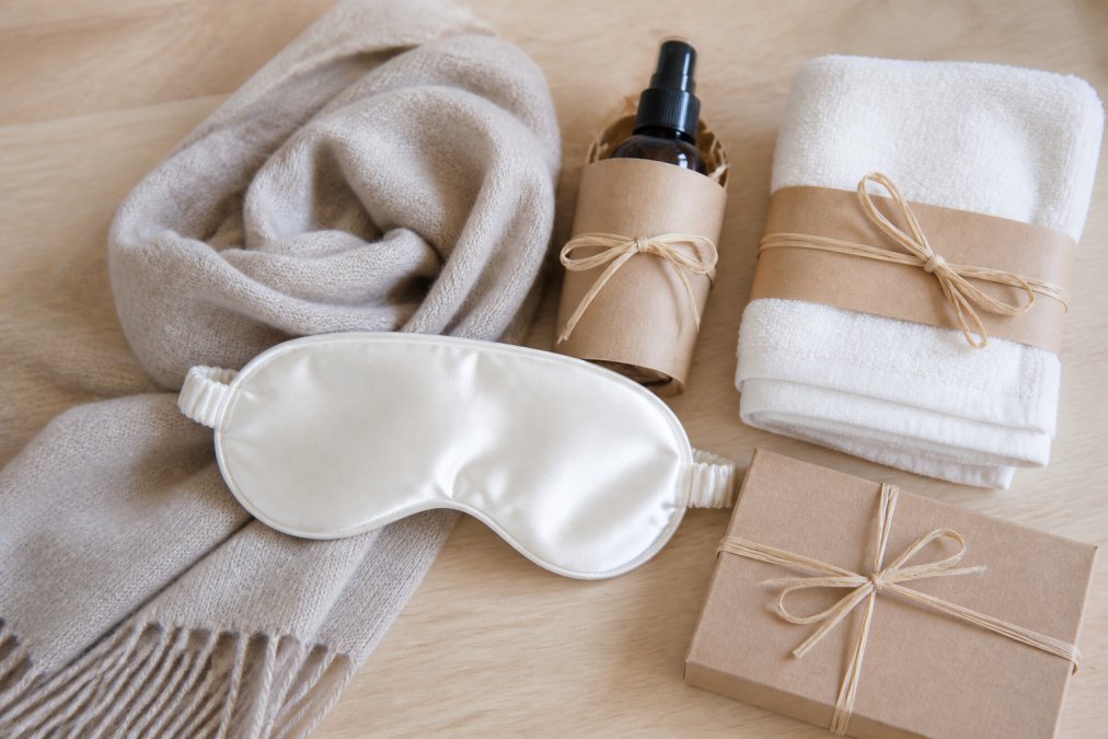 Flat-lay-Fotografie auf einer hellen Holzoberfläche: ein gefalteter beigefarbener Kaschmirschal, eine Seiden-Schlafmaske in Cremeweiß, ein kleines Naturparfum-Fläschchen und ein gefaltetes Baumwollhandtuch, alles in minimalistischer Naturpapier-Verpackung mit Bast-Schleife