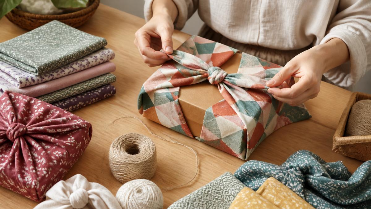 Hände falten ein buntes Baumwolltuch mit geometrischen Mustern um ein rechteckiges Geschenk, mehrere gefaltete Stoffe und Garne liegen auf einem hellen Holztisch verteilt