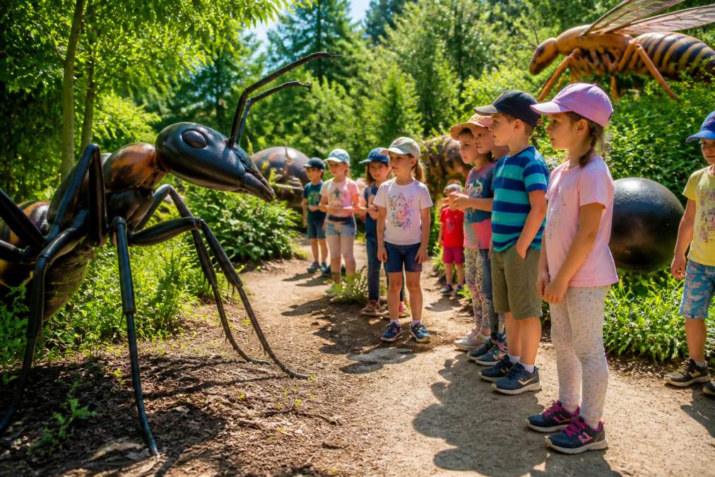 Dzieci w kolorowych ubrankach obserwujące gigantyczne modele owadów na spacerowej ścieżce edukacyjnej w otoczeniu zielonych roślin i drzew, słoneczny dzień