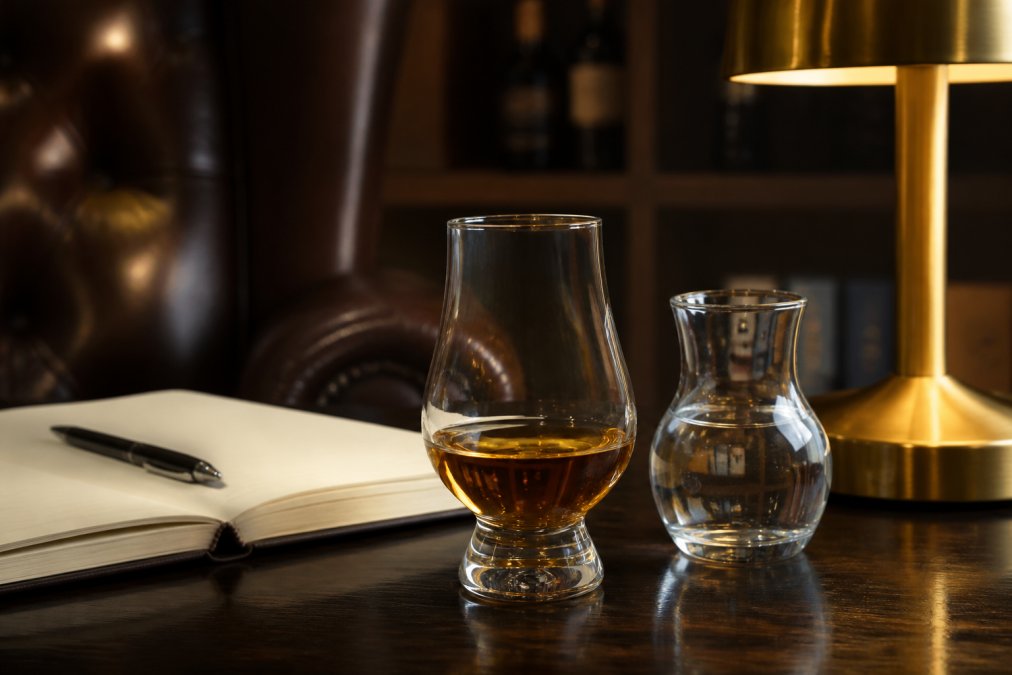 Zbliżenie na kieliszek typu tulipan wypełniony bursztynową whisky, obok stoi mała karafka z wodą źródlaną i otwarty notatnik do degustacji na ciemnym drewnianym stole przy miękkim świetle lampki