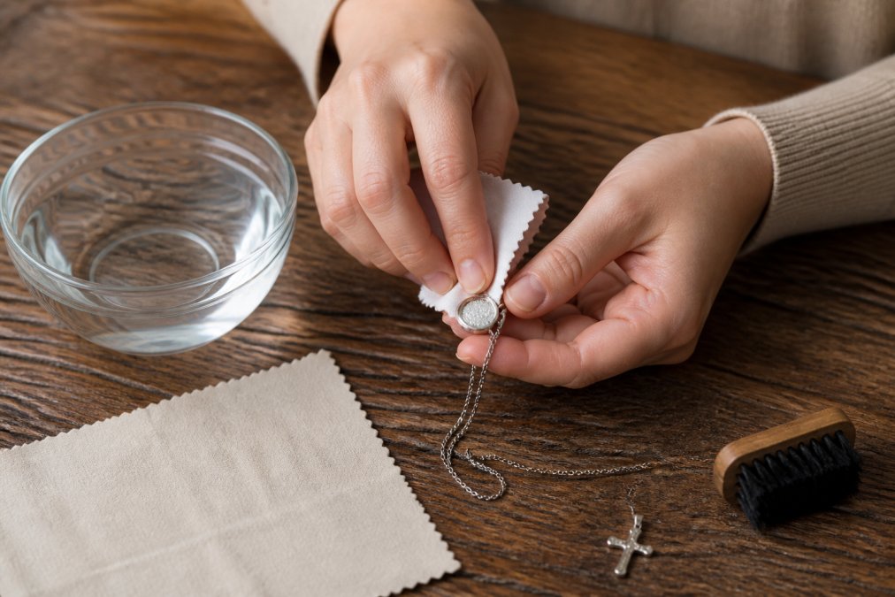 Hands gently cleaning a sterling silver pendant with a soft lint-free cloth on a wooden surface, small bowl of lukewarm water nearby, warm natural lighting
