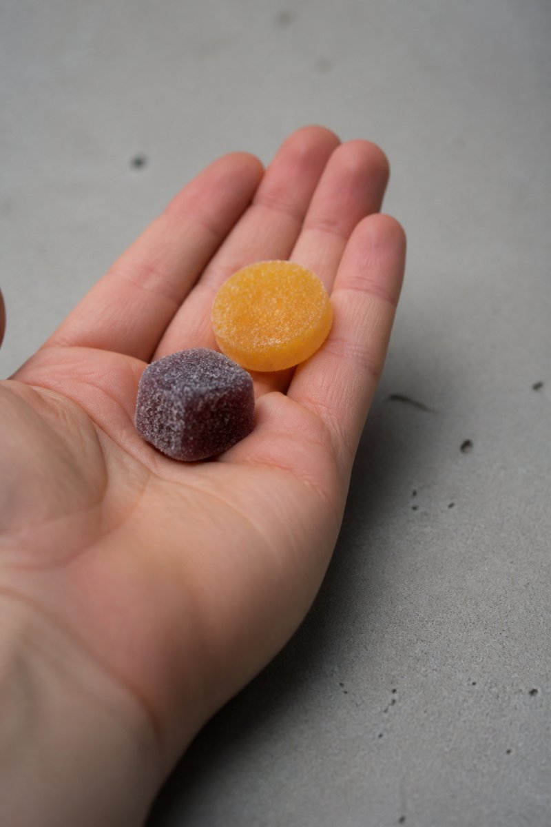 Close-up of a person's hand holding a ZUUZ nanotech tart next to a traditional gummy, with both products clearly visible in natural daylight on a neutral surface
