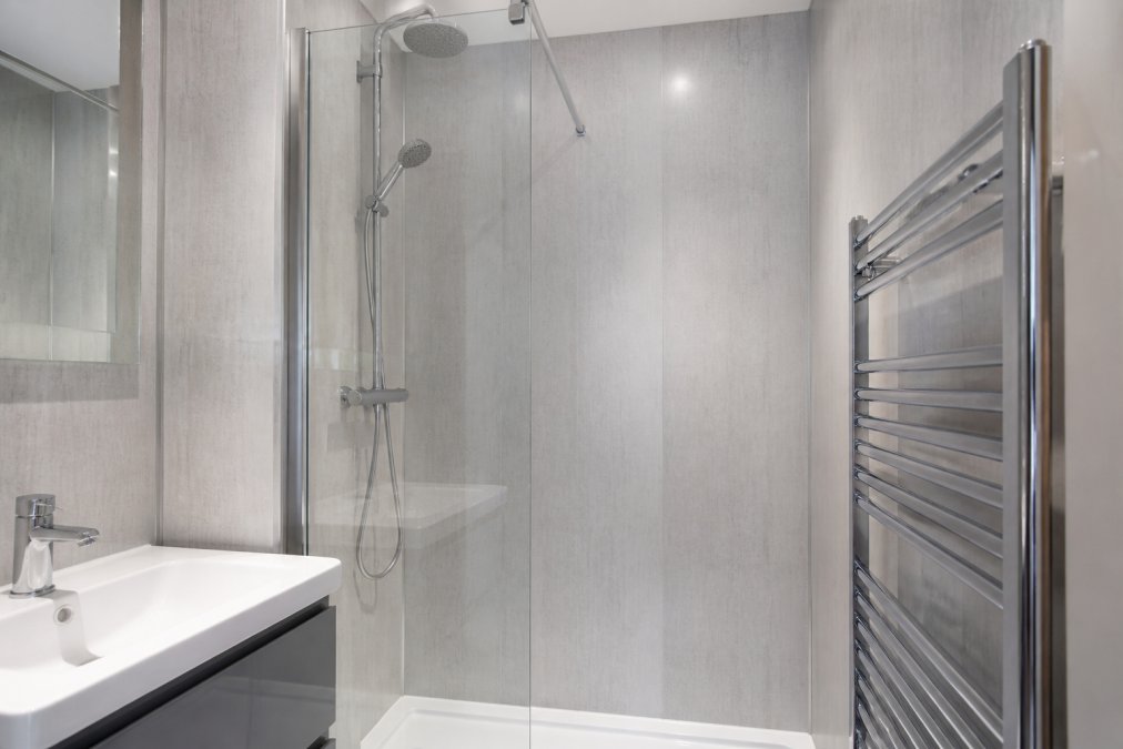 A bright, narrow ensuite shower area with vertical 250mm wall panels in a light grey finish, featuring a glass shower screen and chrome fixtures