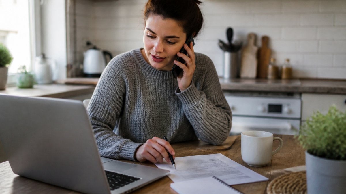 A first-time buyer at a kitchen table with a laptop open, a printed list of questions and a pen, mid-conversation on the phone with a solicitor