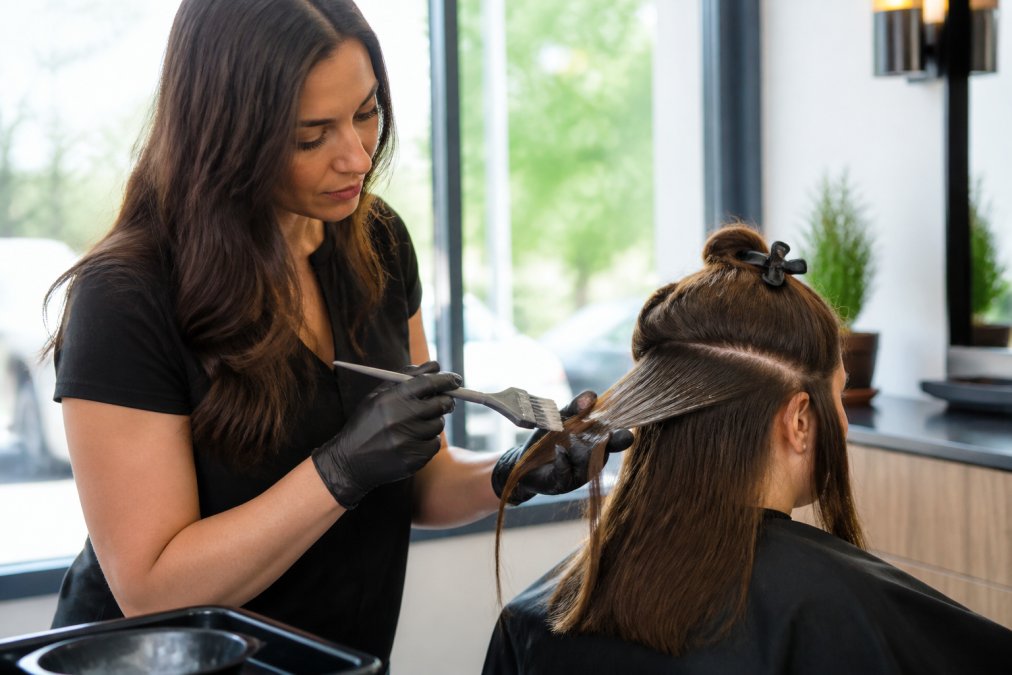 Professional stylist applying keratin treatment to client's hair in a modern salon, showing precise brush application technique on sectioned hair with natural window lighting