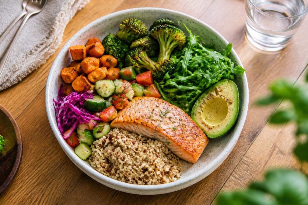 A bright, overhead photograph of a healthy meal bowl filled with leafy greens, colorful vegetables, salmon, and quinoa on a wooden kitchen table, natural window light