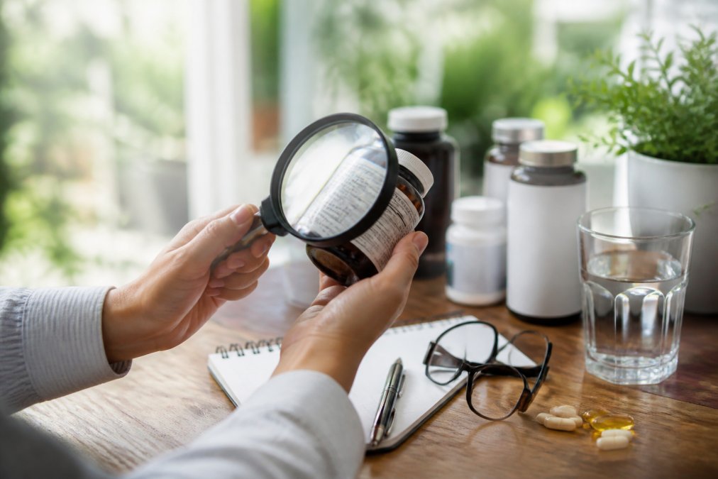 Close-up of hands holding a supplement bottle with a magnifying glass, carefully examining the ingredient label in natural daylight from a window, reading glasses resting on the label