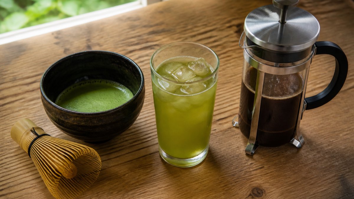 Overhead shot of a wooden table with a matcha whisk and bowl, a glass of iced green tea, and a French press of chicory coffee, natural light from a window