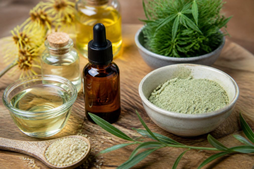 Close-up of natural acne remedy ingredients arranged on a wooden surface with soft natural lighting: tea tree oil in amber bottle with dropper, witch hazel in glass container, hemp seed oil, fresh green clay in ceramic bowl, and sprigs of fresh herbs