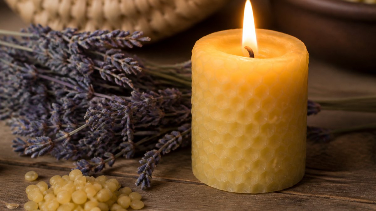 Close-up of a lit beeswax candle with a warm, bright flame and golden glow on a wooden surface next to dried lavender and natural elements, showing the rich cream color and smooth texture of pure beeswax