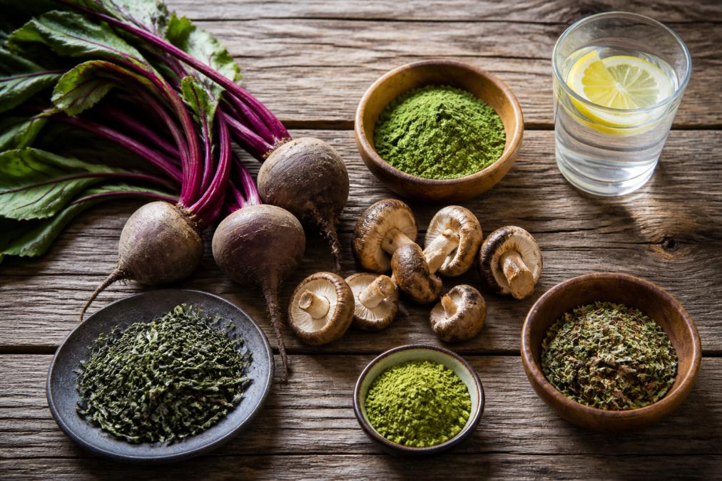 Overhead shot of natural energy ingredients laid out on a wooden surface: fresh beetroot, matcha powder in a bowl, mushrooms, green tea leaves, and a glass of water with lemon
