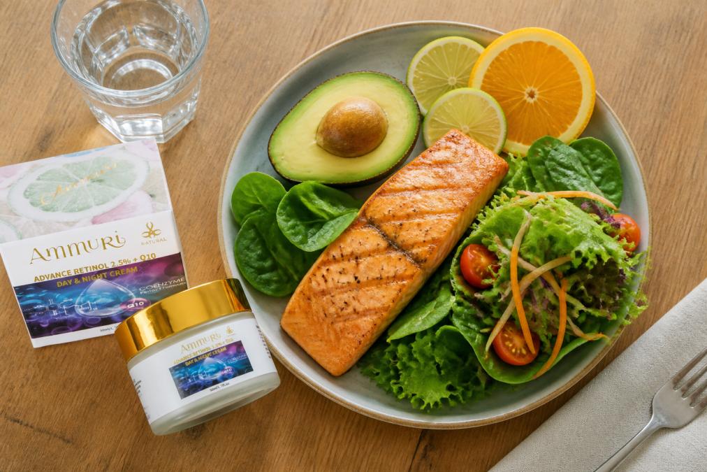 Overhead flat lay of a colourful lunch plate featuring grilled salmon fillet, vibrant leafy greens, fresh citrus slices, and avocado half on a natural wooden table with soft diffused lighting