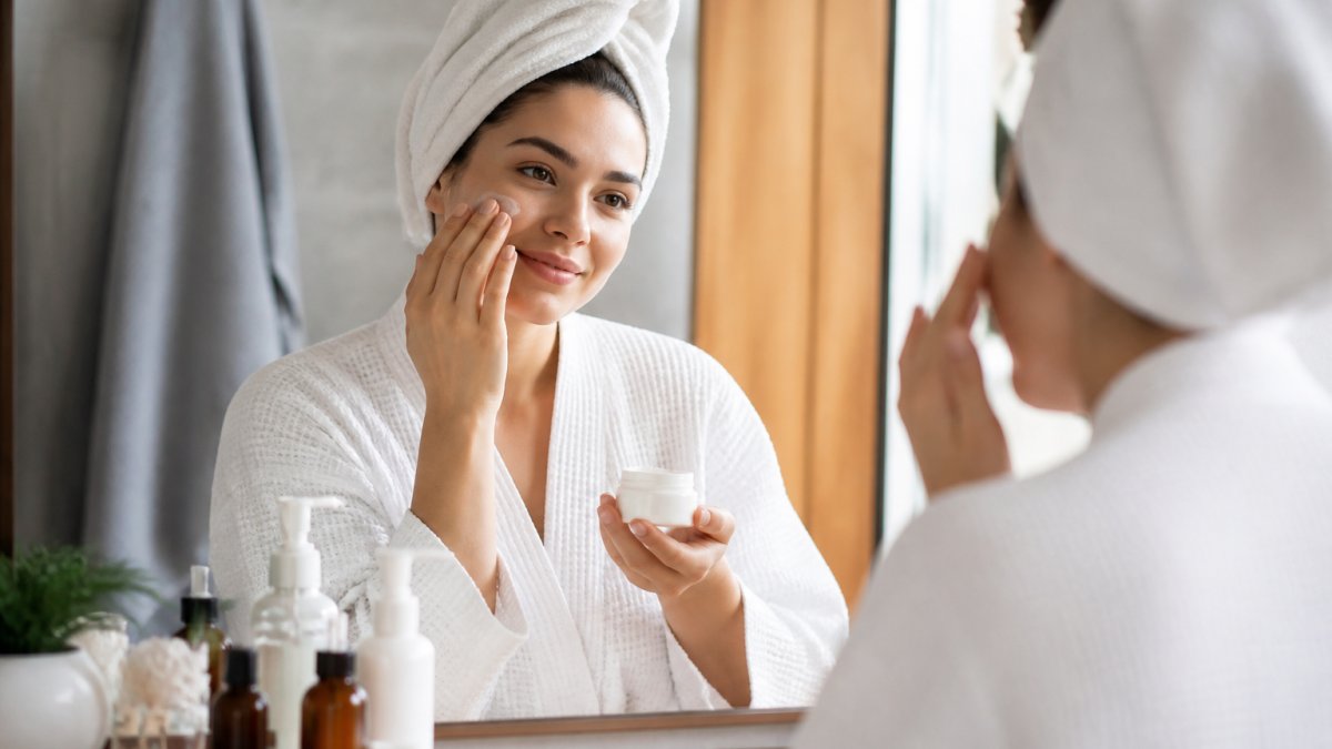 Woman applying moisturizer to her face in a bright bathroom, with skincare products arranged on the counter beside her