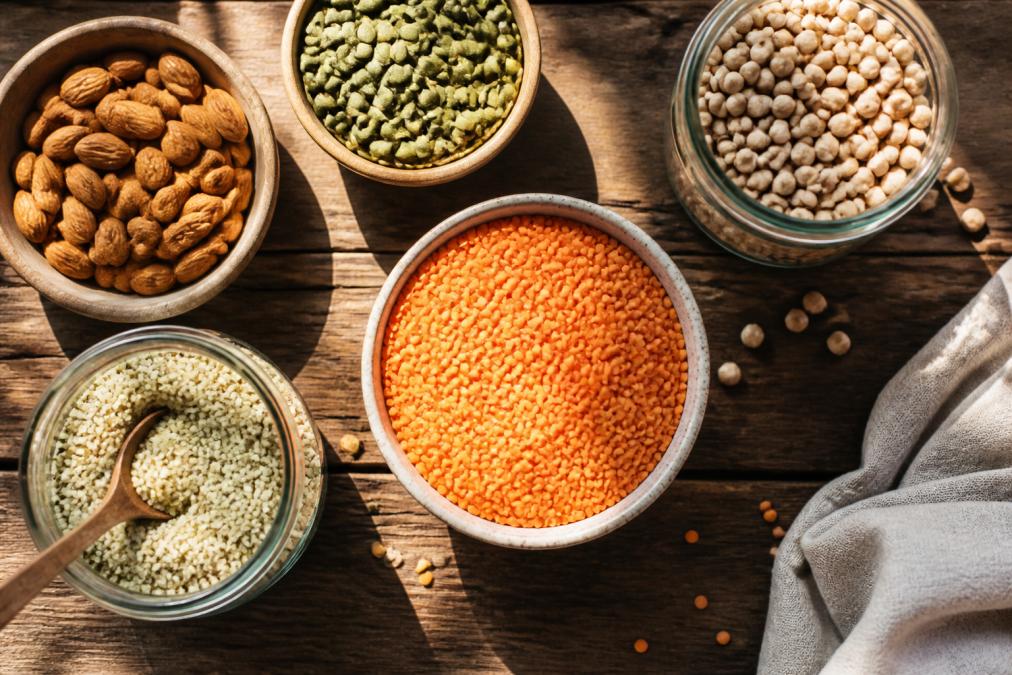 Overhead shot of colorful plant-based foods including lentils, chickpeas, almonds, pumpkin seeds, and hemp seeds arranged on a wooden table with natural sunlight streaming across