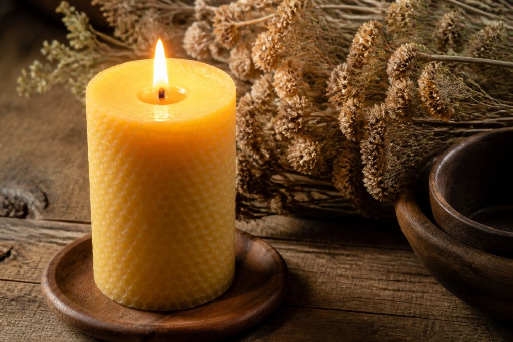 A close-up of a lit beeswax pillar candle on a rustic wooden table, soft golden glow illuminating the surrounding surface, dried flowers in the background
