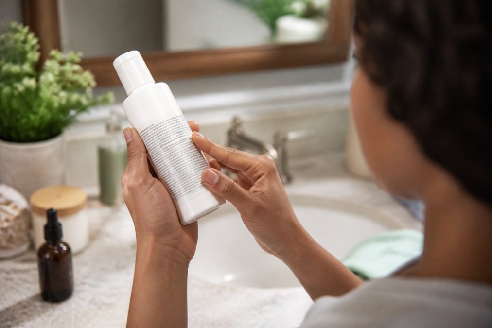 Person in a bright bathroom reading the ingredient list on the back of a skincare bottle
