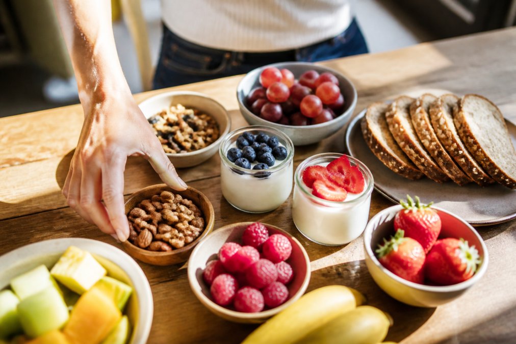 Spread of fresh fruits, nuts, Greek yogurt cups, and whole grain bread arranged on a bright wooden table with natural sunlight streaming across the scene, showing someone's hand reaching for a healthy snack