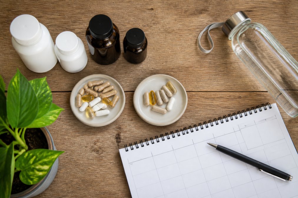 Overhead view of supplement bottles and capsules arranged on a wooden surface with a calendar and water bottle, showing daily routine setup with morning and evening sections clearly separated