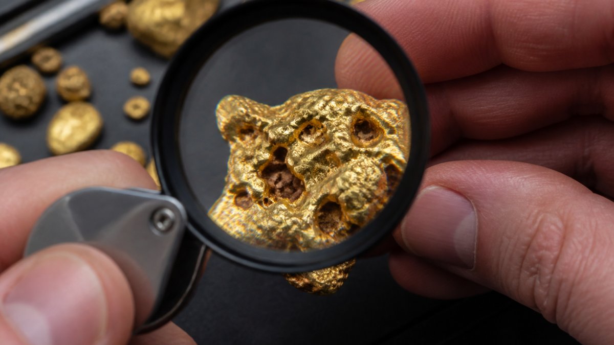 Close-up of a magnifying glass inspecting the organic texture and surface cavities of a natural gold nugget.