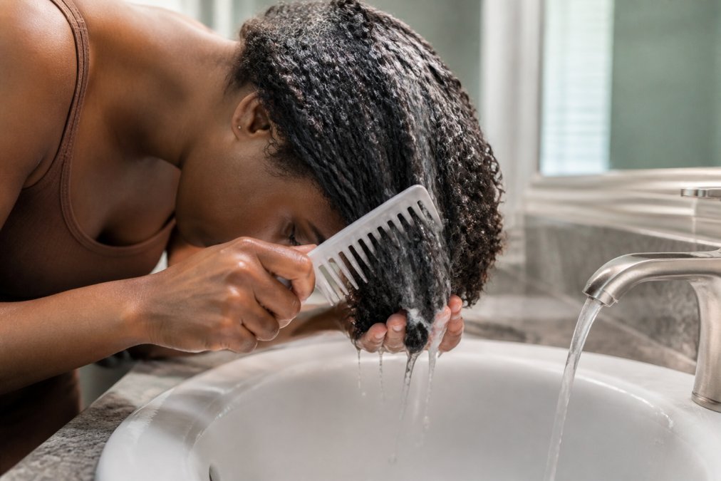 Person with natural curly hair applying conditioner and detangling with a wide-tooth comb over a sink, showing proper technique and water flowing through curls