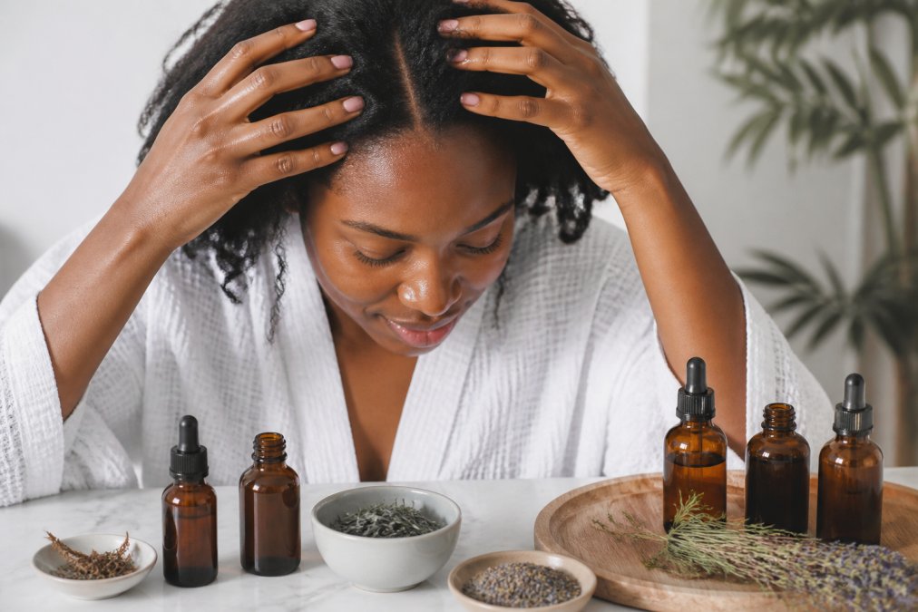 Close-up of a woman's hands gently massaging rosemary and jojoba oil into her scalp, small amber glass dropper bottles and dried herbs arranged on a white marble surface beside her