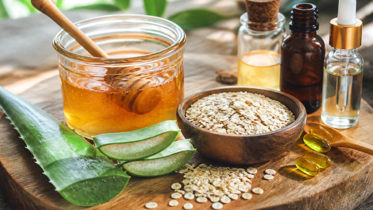 Close-up of natural skincare ingredients arranged on a wooden surface: raw honey in a clear glass jar, aloe vera leaf, rolled oatmeal in a small bowl, and botanical extract bottles with natural lighting
