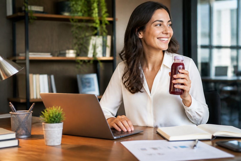 Busy professional at a modern desk holding a fresh natural energy drink bottle, laptop open, appearing focused and energized with natural office lighting