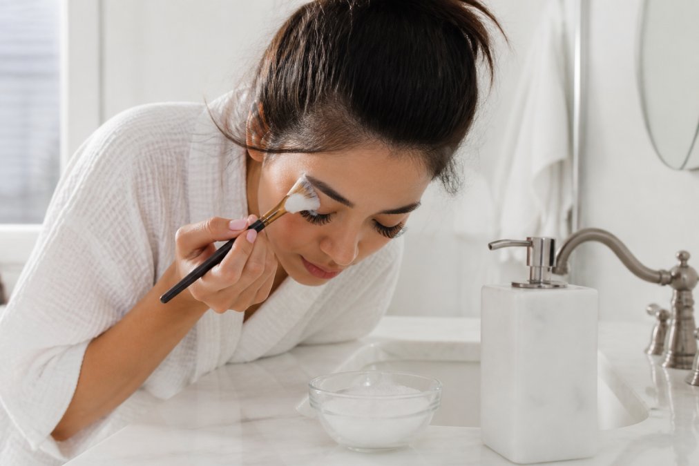 A woman gently cleansing her lash extensions with a soft fan brush and foam cleanser, leaning over a white marble sink in a bright, clean bathroom with natural morning light