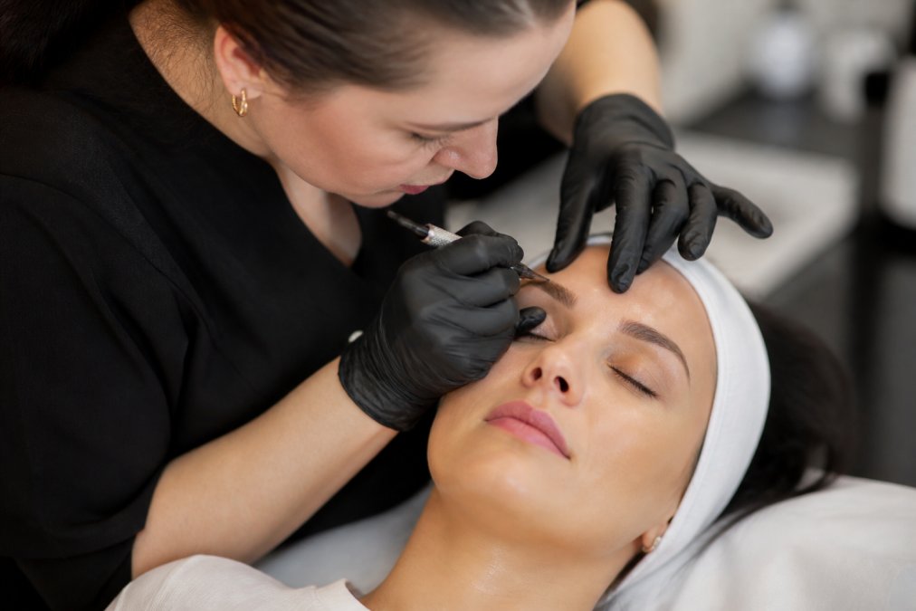 Close-up of a brow artist's hands using a fine manual tool to create delicate hair strokes on a client's eyebrow, with precise pigment application visible under bright clinical lighting
