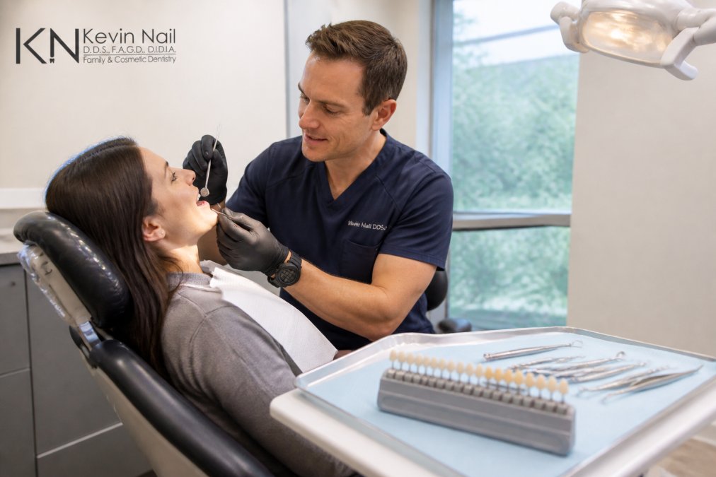 A dentist in gloves carefully examining a patient's teeth in a modern dental chair, dental tools and a shade guide visible on the tray beside them, bright clinical lighting overhead