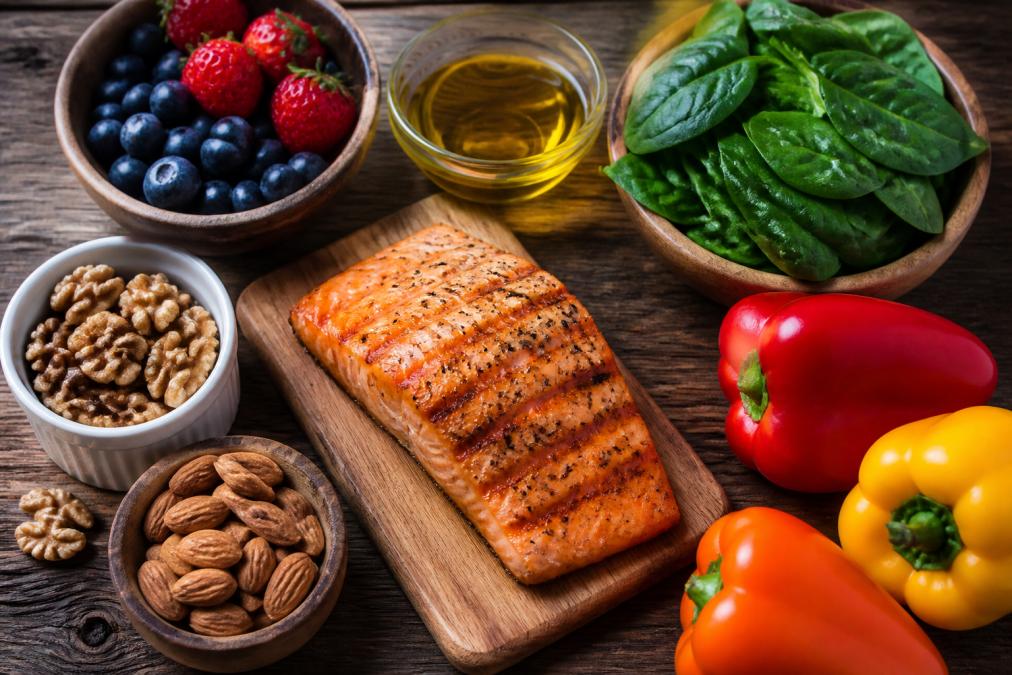 Overhead flat-lay of anti-inflammatory foods arranged on a wooden surface: grilled salmon fillet, fresh blueberries and strawberries, dark leafy spinach, walnuts, almonds, olive oil in a small glass bowl, and colorful bell peppers