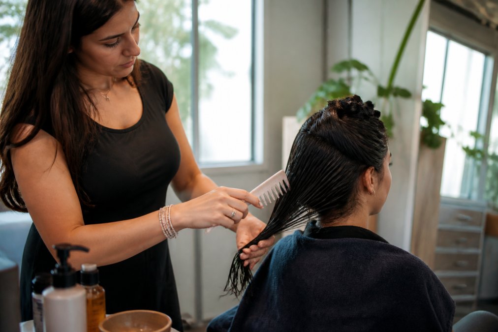 Professional demonstrating oil application technique on damp hair, using a wide-toothed comb to distribute product evenly from mid-shaft to ends in bright, natural lighting