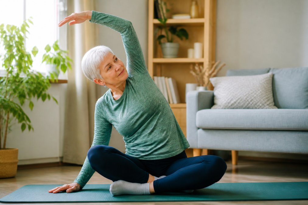 Senior performing gentle stretching exercise in bright living room with natural window light, demonstrating accessible mobility work with proper posture