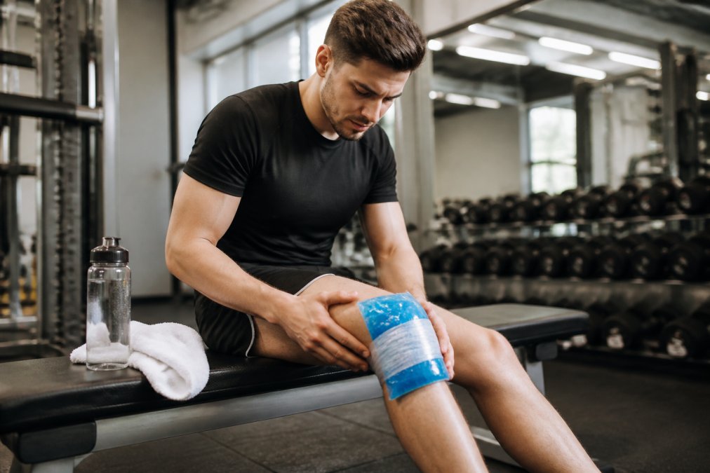 Athlete sitting on a bench with ice pack wrapped around their leg, demonstrating the RICE method, with water bottle and towel nearby in a bright athletic facility