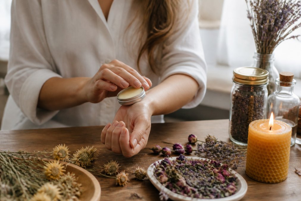 A woman gently dabbing natural solid perfume onto her wrist from a small tin, with dried flowers and a beeswax candle on a wooden table in a bright, airy room